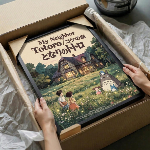 May include: Hands holding a framed vintage-style poster featuring the iconic forest spirit Totoro and two children in a magical woodland scene with a cozy house in the background. The artwork displays warm earth tones and Studio Ghibli's distinctive animation style. The black-framed print is being unpacked from brown cardboard packaging with white tissue paper on a light wooden surface.