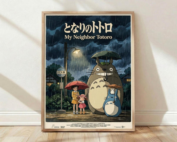 May include: Framed poster of iconic Studio Ghibli characters standing at a rainy bus stop scene, featuring large gray forest creature with umbrella alongside two children under red umbrella, with smaller blue creatures nearby. Dark stormy sky with heavy rain, warm yellow streetlight glow, lush green vegetation. Artwork displayed in light wood frame against white wall on wooden floor with soft natural lighting and shadows.