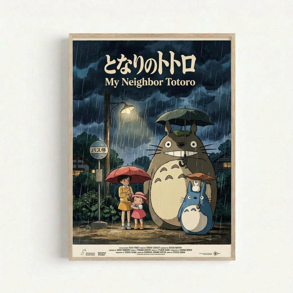 Framed poster featuring the iconic Studio Ghibli film My Neighbor Totoro in a light wood frame against a white wall. The artwork shows two young girls with umbrellas standing at a rainy bus stop next to the large gray Totoro character and smaller blue Totoro, all sheltering from heavy rain under a street lamp. Japanese text appears at the top with English translation below. The scene captures the magical, nostalgic animation style with muted blues and greens depicting the stormy evening atmosphe