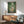 May include: Framed Studio Ghibli My Neighbor Totoro poster displayed above wooden sideboard in modern living room. Artwork shows children with large gray forest creature holding umbrella in lush green bamboo forest. Warm natural lighting illuminates mint green upholstered chair with wooden arms and small table lamp on credenza.