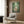May include: Studio Ghibli My Neighbor Totoro movie poster displayed in modern living room setting with beige armchair, geometric throw pillow, brass floor lamp, and dark wood paneling. Poster shows iconic scene of Totoro character with children in lush bamboo forest, featuring green tones and cream frame against warm brown wall.