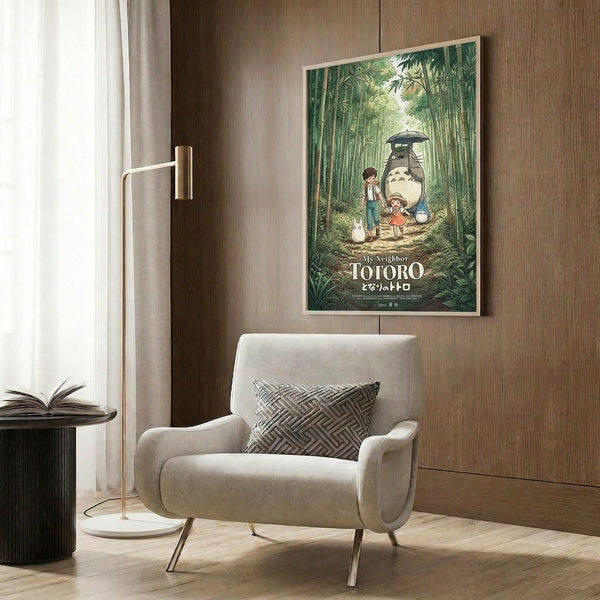 May include: Studio Ghibli My Neighbor Totoro movie poster displayed in modern living room setting with beige armchair, geometric throw pillow, brass floor lamp, and dark wood paneling. Poster shows iconic scene of Totoro character with children in lush bamboo forest, featuring green tones and cream frame against warm brown wall.