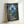 May include: Framed poster in light wood frame leaning against white wall. Artwork shows nighttime forest scene with large Totoro silhouette among moss-covered trees under crescent moon. Small girl in red dress and hat stands on forest path looking up. Fireflies glow throughout dark green foliage. Japanese and English text at bottom. Vintage movie poster aesthetic with blue-green color palette.