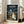 May include: Framed Studio Ghibli My Neighbor Totoro movie poster leaning against white wall next to beige sofa in bright living room. Poster shows iconic rainy bus stop scene with large gray Totoro holding umbrella, two children with red umbrellas, and smaller blue Totoro. Japanese text at top with English title below. Dark stormy sky with street lamp creates atmospheric lighting. Light wood frame complements minimal home decor setting.