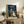 May include: Framed My Neighbor Totoro movie poster in light wood frame displayed on rustic wooden console table alongside fern plant, ceramic vase, and books. Poster shows iconic rainy bus stop scene with Totoro, children under umbrellas, and Japanese text against stormy blue-green sky. Cozy living room setting with beige armchair and neutral wall.