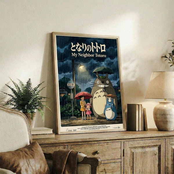 May include: Framed My Neighbor Totoro movie poster in light wood frame displayed on rustic wooden console table alongside fern plant, ceramic vase, and books. Poster shows iconic rainy bus stop scene with Totoro, children under umbrellas, and Japanese text against stormy blue-green sky. Cozy living room setting with beige armchair and neutral wall.
