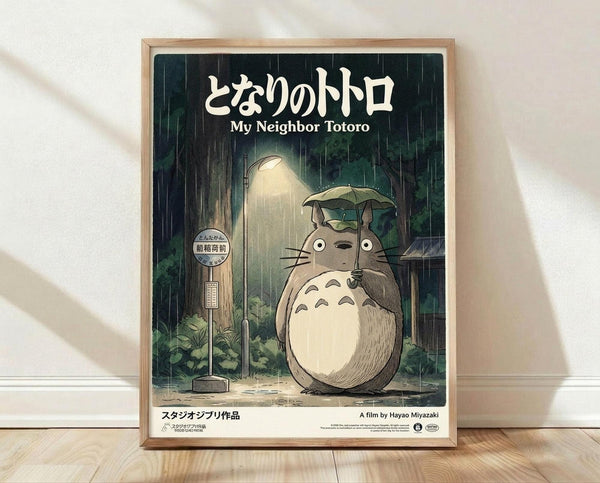 May include: Framed poster print featuring the beloved forest spirit Totoro standing at a rainy bus stop, holding a leaf umbrella while sheltering a small character beneath. The artwork shows Studio Ghibli's distinctive animation style with muted greens and grays depicting heavy rainfall. Japanese text appears at the top with English translation below. The wooden frame sits on light hardwood flooring against a white wall with natural lighting casting shadows.