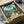 May include: Framed My Neighbor Totoro poster artwork being unpacked from cardboard shipping box with protective wrapping. The colorful illustration shows Totoro characters in a lush green Japanese village setting with traditional houses, mountains, and blue sky. Frame appears black, nestled in brown packaging materials on white tissue paper.