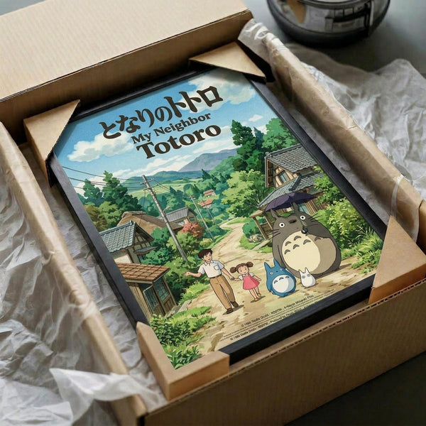 May include: Framed My Neighbor Totoro poster artwork being unpacked from cardboard shipping box with protective wrapping. The colorful illustration shows Totoro characters in a lush green Japanese village setting with traditional houses, mountains, and blue sky. Frame appears black, nestled in brown packaging materials on white tissue paper.