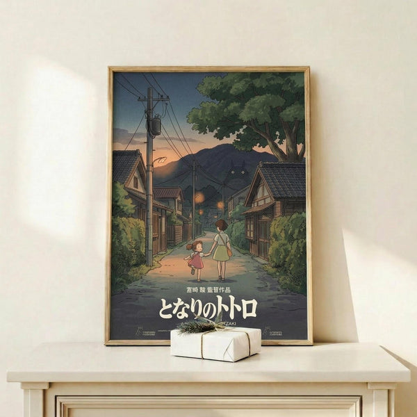 May include: Framed anime-style poster showing two children walking down a rural Japanese village street at dusk, with traditional houses, power lines, mountains, and lush green trees. The artwork features warm orange sunset colors transitioning to deep blue sky. Japanese text appears at bottom. Wooden frame sits on light wood shelf next to small white wrapped gift with natural twine and greenery sprig against cream wall.