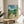 May include: Framed Studio Ghibli My Neighbor Totoro movie poster displayed on a round wooden side table next to a beige upholstered chair. The colorful illustrated poster shows Japanese text, English title, rural village scene with traditional houses, mountains, trees, and iconic Totoro characters on a dirt path under blue sky with white clouds. Natural sunlight streams across the neutral-toned room setting.