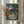 May include: Person holding framed Studio Ghibli My Neighbor Totoro movie poster in light wood frame. Artwork shows two children walking down village street at dusk with traditional Japanese houses, power lines, mountains, and large tree. Warm sunset colors of orange, green, and blue. Japanese text and film credits at bottom. Dried pampas grass visible beside frame in bright, naturally lit room.