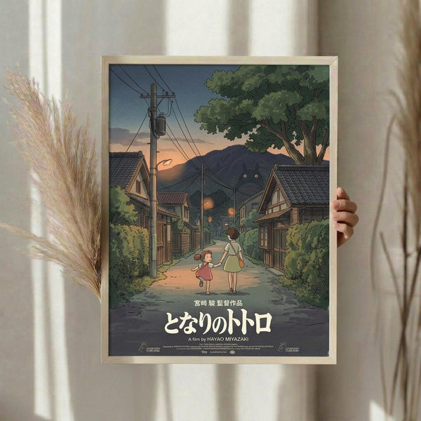 May include: Person holding framed Studio Ghibli My Neighbor Totoro movie poster in light wood frame. Artwork shows two children walking down village street at dusk with traditional Japanese houses, power lines, mountains, and large tree. Warm sunset colors of orange, green, and blue. Japanese text and film credits at bottom. Dried pampas grass visible beside frame in bright, naturally lit room.