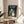 May include: Framed vintage Japanese movie poster featuring beloved forest creature Totoro standing at rainy bus stop with umbrella, displayed on round wooden side table next to beige upholstered chair. Soft natural lighting creates gentle shadows across cream walls, with green plant leaves visible in corner, creating cozy home decor scene.