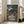 May include: Framed Studio Ghibli movie poster showing two children walking down a traditional Japanese village street at twilight. The anime-style artwork features wooden houses with tiled roofs, power lines, lush green trees, and mountains in the background under an orange-pink sunset sky. Japanese text appears at the bottom. The light wood frame leans against a white wall next to a beige sectional sofa in a bright, minimalist living room with large windows.