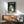 May include: My Neighbor Totoro movie poster in wooden frame hanging above mid-century modern furniture. Shows iconic gray Totoro character holding leaf umbrella in rain at bus stop, with Japanese text at top. Poster displayed in bright living room with teal upholstered chair, wood credenza, white table lamp, and natural sunlight streaming across white walls.