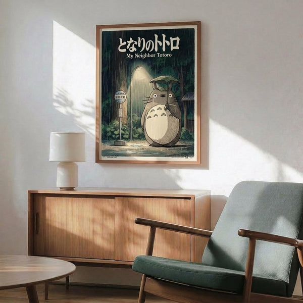 May include: My Neighbor Totoro movie poster in wooden frame hanging above mid-century modern furniture. Shows iconic gray Totoro character holding leaf umbrella in rain at bus stop, with Japanese text at top. Poster displayed in bright living room with teal upholstered chair, wood credenza, white table lamp, and natural sunlight streaming across white walls.