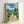 May include: A framed Studio Ghibli My Neighbor Totoro movie poster in a light wood frame, leaning against a white wall. The colorful illustrated poster shows characters including large gray Totoro, smaller blue Totoro, white rabbit, and two children standing on a dirt path through a lush green Japanese village with traditional houses, trees, mountains, and blue sky with white clouds.