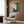 May include: Framed My Neighbor Totoro movie poster hanging on wood-paneled wall above beige upholstered armchair with geometric patterned pillow. Poster features gray Totoro character sitting in lush green forest with Japanese text. Modern reading nook with brass floor lamp, dark round side table with open book, and white curtains visible.