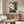 May include: My Neighbor Totoro vintage-style poster mounted on white wall above mid-century modern walnut credenza in minimalist living room. Poster shows gray Totoro character sitting in lush green forest with Japanese text. Room features cream upholstered swivel chair, oval glass coffee table, white table lamp, and wooden floor lamp. Warm natural lighting creates cozy atmosphere with beige walls and contemporary Scandinavian-inspired furniture.