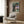 May include: Framed My Neighbor Totoro movie poster displayed on wood-paneled wall above modern beige armchair with geometric pillow. Poster shows iconic characters under large tree at twilight with Catbus in branches. Contemporary living room setting with brass floor lamp and side table.