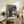 May include: Framed vintage-style Studio Ghibli poster displayed on rustic wooden sideboard in cozy living room setting. Poster shows Totoro characters under large tree with Catbus in branches, rendered in muted earth tones against twilight sky. Scene includes fern plant, ceramic vase, stacked books, and cream-colored armchair with brown throw pillows.