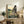 May include: Framed My Neighbor Totoro poster featuring a small child and large gray forest spirit sitting together on a wooden bench under an umbrella, with traditional Japanese house and rolling green countryside in background. Warm sunset colors throughout the artwork. Light wood frame displayed on rustic wooden sideboard alongside fern plant, ceramic vase, books, and beige armchair visible in cozy living room setting with cream walls.