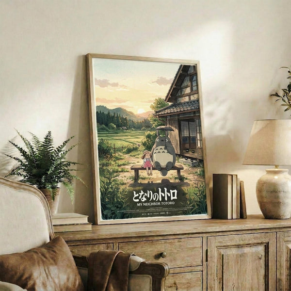 May include: Framed My Neighbor Totoro poster featuring a small child and large gray forest spirit sitting together on a wooden bench under an umbrella, with traditional Japanese house and rolling green countryside in background. Warm sunset colors throughout the artwork. Light wood frame displayed on rustic wooden sideboard alongside fern plant, ceramic vase, books, and beige armchair visible in cozy living room setting with cream walls.
