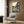 May include: Framed My Neighbor Totoro poster displayed on wood-paneled wall above beige armchair with geometric pillow. Shows Totoro and small girl sitting on wooden bench outside traditional Japanese house at sunset with mountains and fields in background. Japanese text at bottom. Modern living room setting with gold floor lamp and dark round side table.