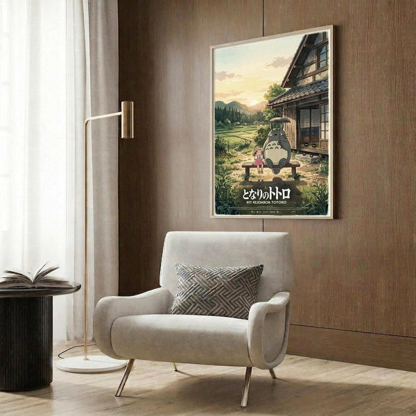 May include: Framed My Neighbor Totoro poster displayed on wood-paneled wall above beige armchair with geometric pillow. Shows Totoro and small girl sitting on wooden bench outside traditional Japanese house at sunset with mountains and fields in background. Japanese text at bottom. Modern living room setting with gold floor lamp and dark round side table.