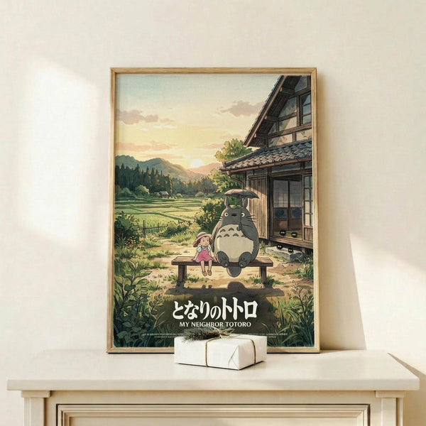 May include: Framed poster showing animated characters Totoro and a young girl sitting on a wooden bench outside a traditional Japanese house, with rolling hills and rice fields in the background during golden hour. The artwork features soft, warm colors in Studio Ghibli's distinctive animation style. Japanese text and English title appear at bottom. Light wood frame sits on white wooden surface next to small white gift box against cream wall.