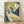 May include: Framed Art Nouveau poster print featuring an elegant woman with flowing red hair in a cream dress with floral blue sleeves, reading a newspaper. She sits in an ornate green chair with decorative stained glass-style background in greens and golds. The vintage illustration shows "International Studio" text at bottom with artist's monogram. Natural wood frame displayed against white wall on light wooden floor with soft shadows.