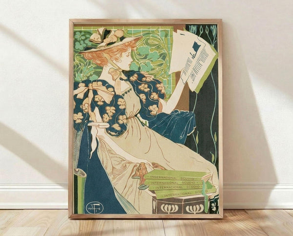 May include: Framed Art Nouveau poster print featuring an elegant woman with flowing red hair in a cream dress with floral blue sleeves, reading a newspaper. She sits in an ornate green chair with decorative stained glass-style background in greens and golds. The vintage illustration shows "International Studio" text at bottom with artist's monogram. Natural wood frame displayed against white wall on light wooden floor with soft shadows.