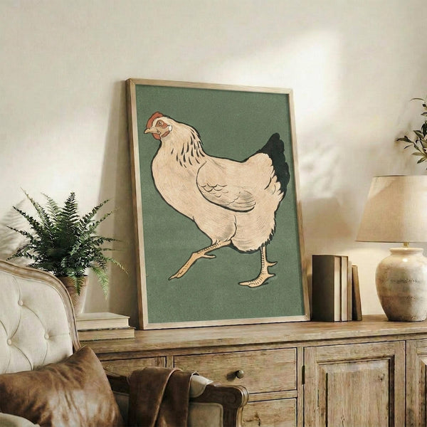May include: Framed vintage Japanese woodblock print of a white hen with red comb walking on sage green background, displayed on rustic wooden sideboard alongside fern plant, ceramic vase, and books in cozy living room setting with cream upholstered chair and warm lighting