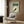 May include: Vintage-style Japanese poster with green background featuring black goldfish silhouette surrounded by concentric circles, displayed in light wood frame on wood paneled wall above beige upholstered chair with geometric patterned pillow, next to brass floor lamp and dark round side table with open book