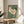 May include: A framed vintage Japanese woodblock print of a white hen with red comb walking against sage green background, displayed on a round wooden side table next to a beige upholstered chair. Natural sunlight streams across the neutral-toned room setting with a plant visible in the corner.