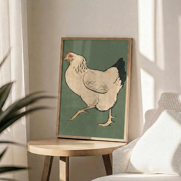 May include: A framed vintage Japanese woodblock print of a white hen with red comb walking against sage green background, displayed on a round wooden side table next to a beige upholstered chair. Natural sunlight streams across the neutral-toned room setting with a plant visible in the corner.