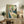 May include: Vintage Art Nouveau poster in light wood frame showing woman in green hat with flowers reading papers, displayed on rustic wooden sideboard with books, ceramic vases, and fern plants in neutral-toned living room setting