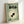 May include: Framed poster with sage green background featuring Japanese text at top, black silhouette of a goldfish with large round eyes in center surrounded by cream-colored concentric circles, small cartoon face illustration in bottom left corner, displayed in light wooden frame leaning against white wall