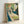 May include: Framed vintage Art Nouveau poster print leaning against white wall, featuring elegant woman with flowing red hair in floral blue dress holding papers, surrounded by green foliage and books, displayed in light wood frame on white surface.
