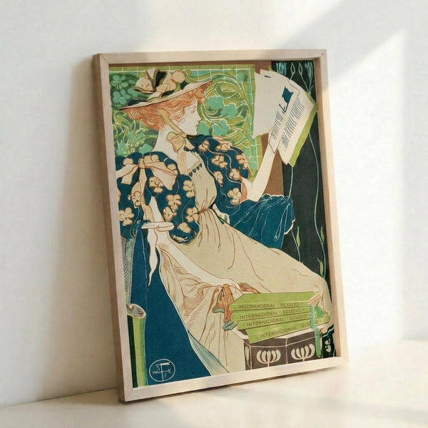 May include: Framed vintage Art Nouveau poster print leaning against white wall, featuring elegant woman with flowing red hair in floral blue dress holding papers, surrounded by green foliage and books, displayed in light wood frame on white surface.