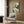 May include: Art Nouveau style poster featuring woman with flowing green hair in white dress, displayed in wooden frame on wood-paneled wall above beige upholstered armchair with geometric pillow. Modern brass floor lamp and dark round side table with open book complete the sophisticated reading nook scene with natural lighting from window.