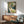 May include: Vintage Art Nouveau poster print with elegant woman in flowing white dress and floral crown, displayed in thin wood frame above mid-century modern credenza. Room features sage green upholstered chair with wooden arms, white ceramic table lamp, and warm sunlight casting shadows on white wall. Poster shows muted green, cream and blue color palette in decorative Victorian advertising style.