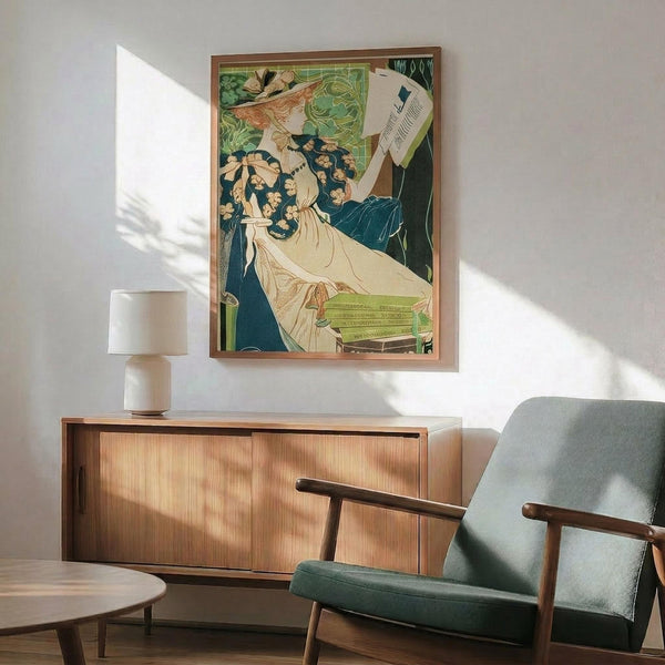 May include: Vintage Art Nouveau poster print with elegant woman in flowing white dress and floral crown, displayed in thin wood frame above mid-century modern credenza. Room features sage green upholstered chair with wooden arms, white ceramic table lamp, and warm sunlight casting shadows on white wall. Poster shows muted green, cream and blue color palette in decorative Victorian advertising style.