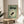 May include: Framed Japanese-style poster with green background featuring black goldfish silhouette in center surrounded by cream-colored wavy rings, Japanese text at top, displayed on light wood round side table next to beige upholstered chair, plant leaves visible in soft natural lighting