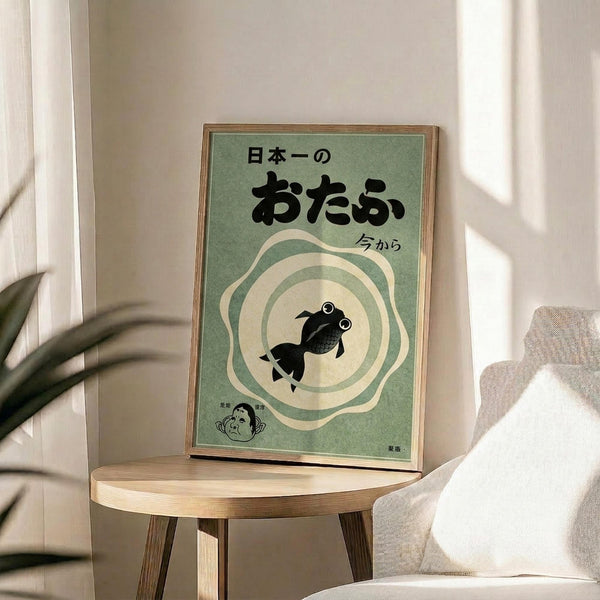 May include: Framed Japanese-style poster with green background featuring black goldfish silhouette in center surrounded by cream-colored wavy rings, Japanese text at top, displayed on light wood round side table next to beige upholstered chair, plant leaves visible in soft natural lighting