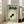 May include: Framed vintage-style Japanese poster with sage green background featuring black goldfish in cream circular ripples, Japanese text at top, small character illustration in bottom corner, displayed on wooden floor next to beige upholstered chair in bright room with white walls and windows.
