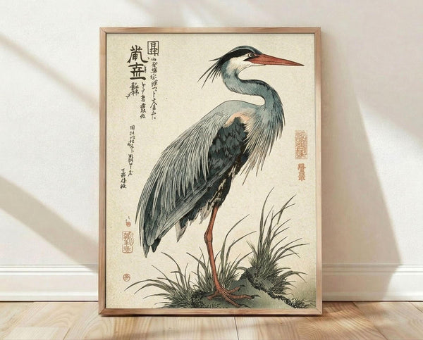 May include: Japanese woodblock print of a grey heron with long orange beak and legs, standing among marsh grasses. The bird displays detailed blue-grey and white plumage with black wing markings. Traditional calligraphy and red seals appear on cream background. Displayed in thin wooden frame on light hardwood floor against white wall with soft natural lighting.