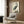 May include: Japanese woodblock print of a grey heron with orange beak standing among reeds, displayed in light wooden frame on dark wood paneled wall above beige upholstered armchair with geometric patterned pillow, brass floor lamp and side table with open book nearby