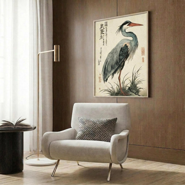 May include: Japanese woodblock print of a grey heron with orange beak standing among reeds, displayed in light wooden frame on dark wood paneled wall above beige upholstered armchair with geometric patterned pillow, brass floor lamp and side table with open book nearby